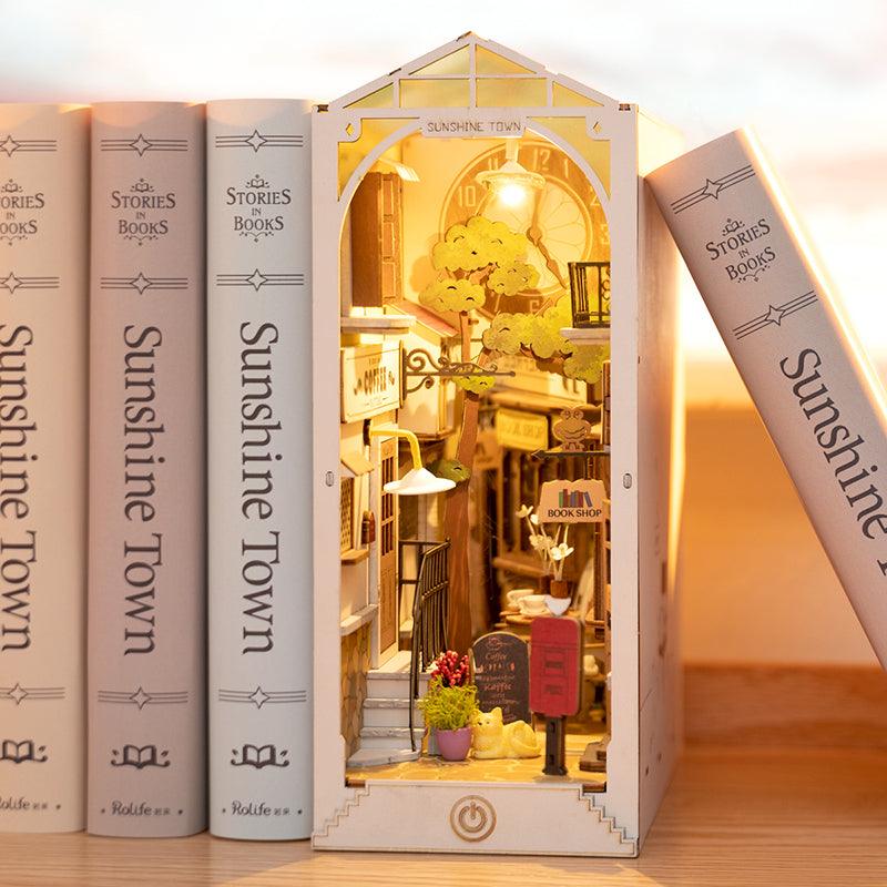 The adorable book nook is shown here how it would be within your own home. It is slotted between some books to demonstrate how it can create this tiny universe within your book shelf. It is shwon with a coffee shop, book shop, tree, illuminated lights and a table with a cup of tiny cup of coffee set upon it. The item is perfect as a reading themed gift for any book lovers.