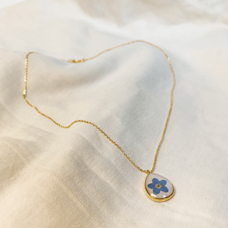 The necklace is laid out on a soft cotton sheet to show it's full beauty. The 14ct gold plated chain has the pendent hanging on one side, and a lobster claw clasp on the other. The pendent is a tear drop shape with a milky white colour and a forget me not in the middle. The pendent is rounded with 14ct gold plating to define it's delicate shape. Jane Austen Jewellery Regency Jewellery 
