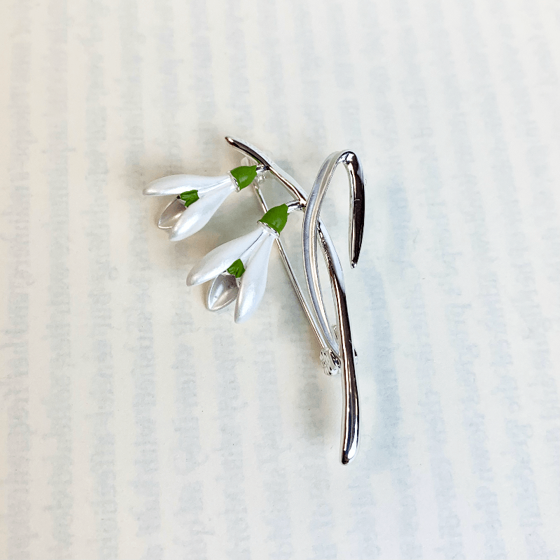 the snowdrop brooch on a white background showing the faint outlines of a book page. Reminiscent of the pages of pride and prejudice which talk about the lovely grounds of Mr Darcy's pemberely